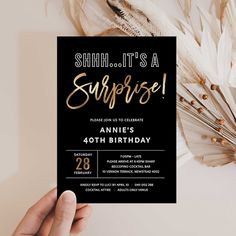 someone holding up a black and gold surprise birthday party card with feathers on the table