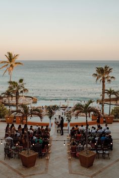 people are sitting at tables in front of the ocean with palm trees on either side