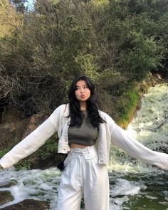 a woman standing in front of a river with her arms spread out and wearing white pants