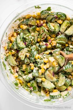 a glass bowl filled with cucumber and corn salad next to a wooden spoon