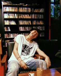 a man sitting on a couch in front of a book shelf filled with books and drinks