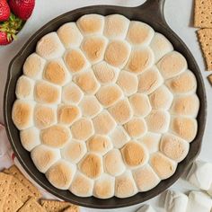 a skillet filled with marshmallows on top of a table next to crackers and strawberries