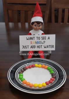 an elf is sitting next to a plate with candy on it and a sign that says, want to see a rainbow just add hot water