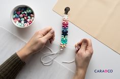 two hands are stringing beads together on a white surface with a cup of coffee in the background