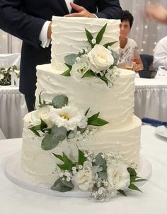 a wedding cake with white flowers and greenery on the top is surrounded by people