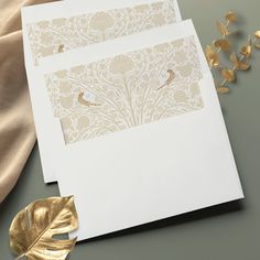 two white cards with gold foiling on them next to a leaf and flower arrangement