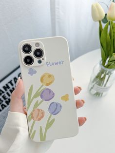 a person holding a phone case with flowers painted on the front and back cover, in front of a vase filled with tulips