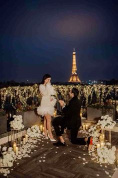 a man kneeling down next to a woman at a table with candles and flowers in front of the eiffel tower