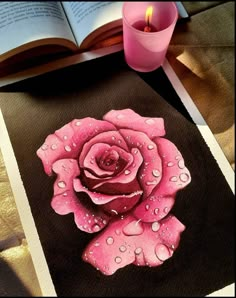 a pink rose with water droplets on it next to an open book and candle holder