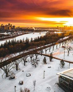 the sun is setting over a snowy park with trees and buildings in the foreground