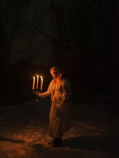 a woman holding three lit candles in the dark with snow on the ground behind her