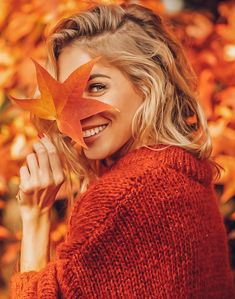 a woman wearing a red sweater and holding an orange maple leaf in front of her face