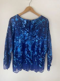 The captivating Blue Sequin Blouse -- designed to mesmerize and elevate your look to new heights. This stunning blouse, in good vintage condition, boasts a few small discolorations on the sequins located on the upper rear neckline. However, rest assured that these minor imperfections do not detract from the overall beauty of this eye-catching piece. Crafted in size L, this blouse is intentionally oversized, delivering a relaxed and comfortable fit for everyone. The sheer fabric and intricate hand-beaded detailing on the lower shirt body and sleeves exude an air of elegance and sophistication. The impeccable condition of the beads and sequins ensures that they catch the light, creating a mesmerizing sparkle wherever you go. Prepare to turn heads and make a stylish statement with the Blue Se Glamorous Blue Tops For Party Season, Blue Sequined Blouse For Spring, Glamorous Blue Sequined Tops, Blue Embellished Long Sleeve Top, Festive Blue Sequined Blouse, Blue Embellished Party Blouse, Festive Blue Sequin Blouse, Blue Sequin Top For Party Season, Blue Sequin Tops For Party Season