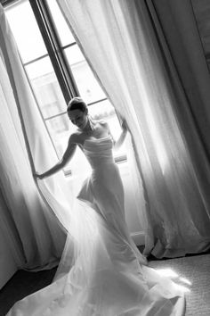 a woman standing in front of a window wearing a white dress and holding her arms out