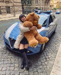 a woman sitting on the hood of a car holding a large teddy bear in front of her