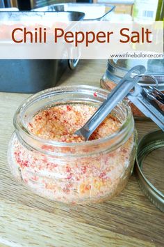 a jar filled with chili pepper salt sitting on top of a wooden table next to a spoon