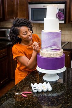 a woman in an orange shirt is decorating a purple and white tiered cake