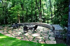 a stone patio surrounded by green grass and trees with chairs around the fire pit in the middle