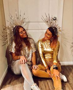 two women sitting on the floor wearing gold outfits and silver headpieces, one with her legs crossed