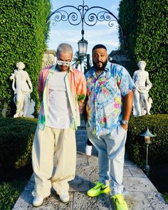 two men standing next to each other in front of some bushes and trees with statues behind them