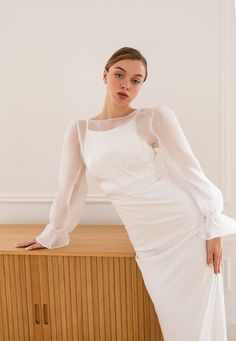 a woman in a white dress is leaning on a wooden cabinet and posing for the camera