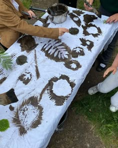 three people are making art on a white table cloth with brown and green leaves in the background