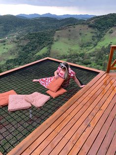 a woman laying on top of a wooden deck next to a lush green hillside covered in trees