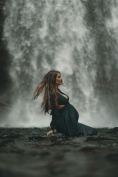 a woman sitting in front of a waterfall with her hair blowing in the wind and flowing water behind her