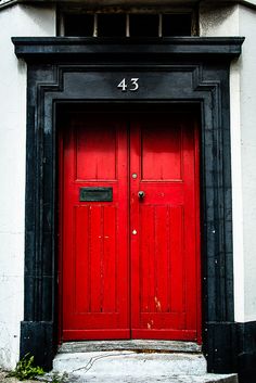 a red door is on the side of a white building with black trim and numbers