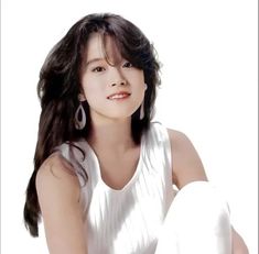 a woman with long dark hair sitting on the ground wearing white clothing and earrings, looking at the camera