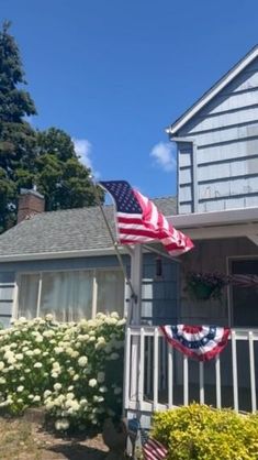 an american flag on the front porch of a house with white flowers and bushes around it