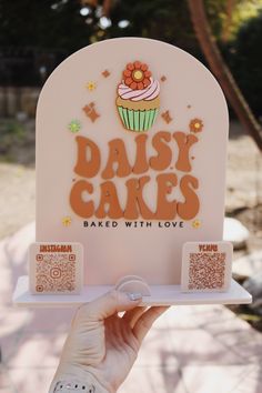 a person holding up a sign that says daisy cakes baked with love and cupcakes on it