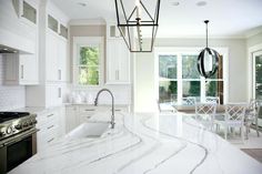 a white kitchen with marble counter tops and an island in front of a window that looks out onto the backyard