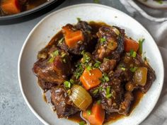 a white plate topped with beef and carrots next to a bowl of soup on a table