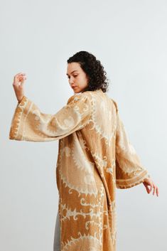 This semi-fitted duster was made from an antique hand-embroidered woven cotton Suzani, a textile traditionally used as wall hangings or in the yurts of nomadic people in Central Asia. An original aloka design, we prepared the vintage material with our signature multi-step shampoo and softening process. In our Athens, Georgia-based studio, our designers created this A-line semi fitted jacket with a plunging neckline and puffed bell sleeves. Served bound seams on the interior showcase the hand-stitching. Thick trim along the bottom of the jacket and sleeves give it a luxurious look and drape. Each textile bares memories and marks of the hand and past. Each piece is a one-of-a-kind garment. We carefully curate and prepare these beautifully preserved textiles and embrace imperfections, mending Traditional Cotton Ikat Print Kimono, Traditional Cotton Kimono With Ikat Print, Traditional Beige Kaftan For Festivals, Bohemian Embroidered Beige Outerwear, Traditional Long Sleeve Kimono With Ikat Print, Traditional Long Sleeve Ikat Print Kimono, Traditional Cream Kimono, Embroidered Beige Bohemian Kaftan, Traditional Long Sleeve Kimono With Natural Dye