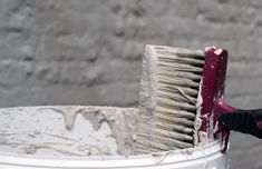 a close up of a brush in a bucket with paint on the outside and brick wall behind it