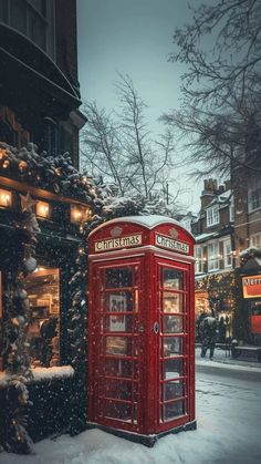 a red phone booth sitting in the middle of a snow covered street next to a christmas tree