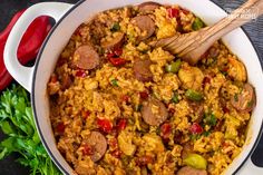 a pot filled with rice, sausage and peppers on top of a table next to a wooden spoon