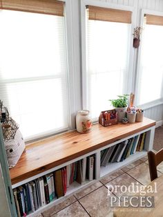 a wooden table topped with lots of books next to two windows covered in blind shades