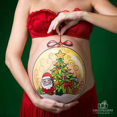 a pregnant woman wearing santa claus's christmas tree tattoo on her belly, while holding an ornament