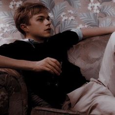 a young man sitting on top of a couch in front of a floral wallpaper