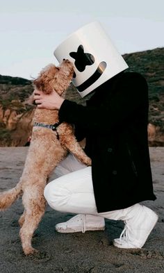 a man holding a dog and wearing a mask on his face while kneeling down in the sand