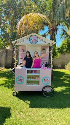 two women standing in a pink and white ice cream cart on grass with palm trees behind them