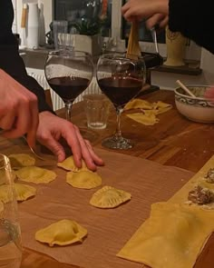 two people are making ravioli on a wooden table with wine glasses and other dishes in the background