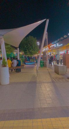 people are walking around an outdoor shopping area at night with umbrellas and benches in the foreground