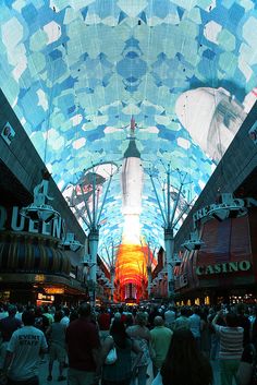 people are walking through an indoor shopping mall in the evening time, with colorful lights on the ceiling