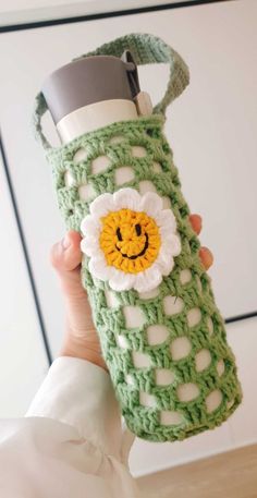 a woman holding up a green crocheted water bottle with a smiling face on it