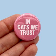 a pink button with the words in cats we trust on it is held in a hand