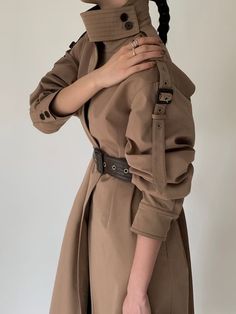 a woman wearing a trench coat and holding her hand on her shoulder with both hands