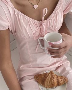 a woman in a pink dress is holding a cup and plate with a croissant on it